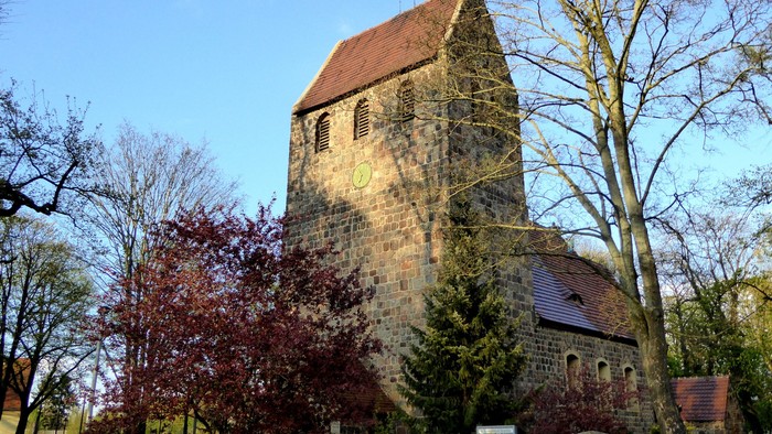 Alte Kirche mit hohem Turm und rotem Dach, umgeben von Bäumen und Büschen