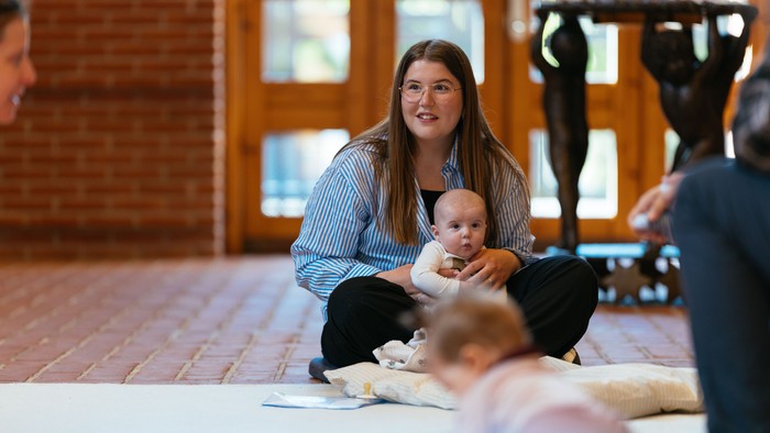 En kvinde sidder på gulv med et barn på skødet, mens et andet barn spiller med legetøj ved siden af.
