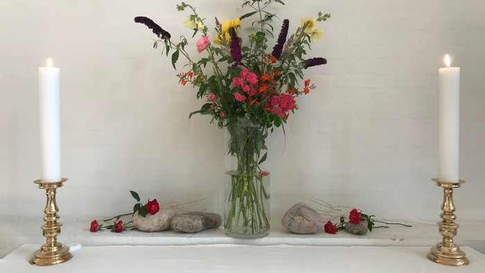En blomsterstang i glasvas med farverige blomster og grønne blade placeret på en hvid bord med to lyse lysestage på hver side.