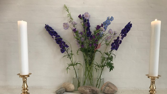 En hvid bordsduk med to gyldne lysestage med lyse lygter, en blomstervase med blå og violette blomster og en åben bog.