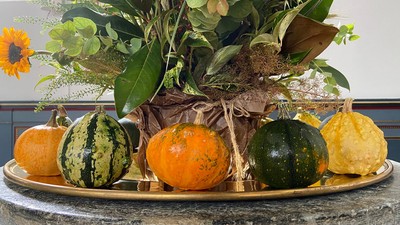 En hyggelig høstdekoration med blomster og grønne planter på en fat og en plate med diverse pumpker og grønsager.