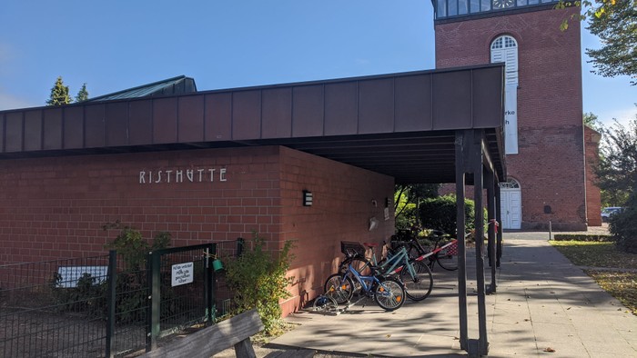 Fahrradunterstand in der Nähe eines Ziegelgebäudes mit der Aufschrift „Risthytte“ und einem Kirchturm im Hintergrund.