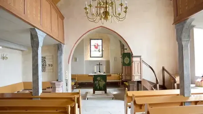 Kleine, ruhige Kircheninnenraum mit hölzernen Kirchenbänken, Kronleuchter und Altar.