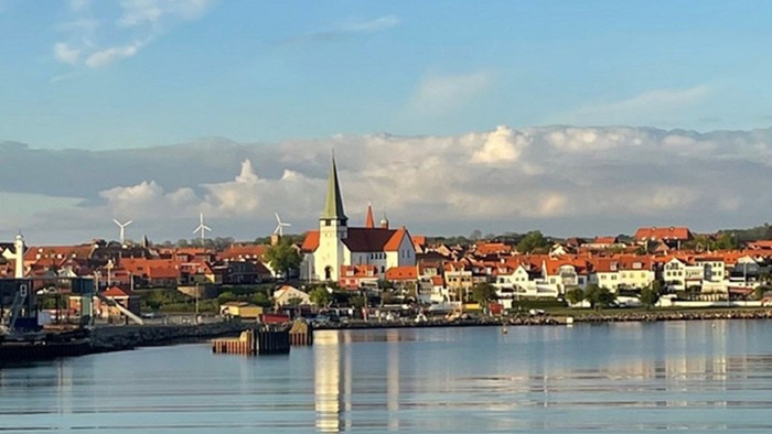En kystby med røde tagtegn og en kirke med spiret ud mod havnen og bjerge i baggrunden