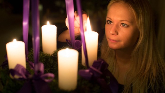En ung pige med længe blond hår holder en adventskranse med lysende lygter og violette bænder.
