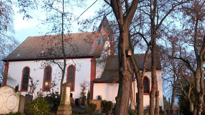 Kleine Kirche mit hohem Kirchturm, umgeben von Bäumen und Gräbern
