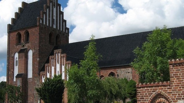 Et stort, gotisk kirkebygning med høje tårne og mure af røde teglsten, omgivet af grønne træer og et klart blå himmel.