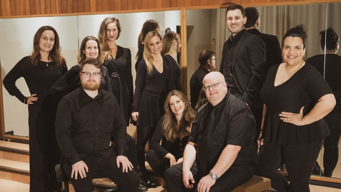 Eine Gruppe von Darstellern in schwarzer Kleidung, die gemeinsam in einem Proberaum oder Aufnahmestudio posieren.