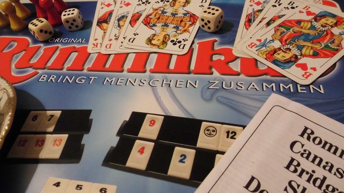 Eine Tischansicht des Kartenspiels Rommé mit Würfeln, Karten, Zählsteinen und Spielregeln.