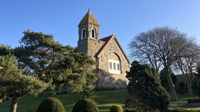 Hyggelig stenkirke med klokketårn omgivet af veltrimmet grønne områder under en klar blå himmel.