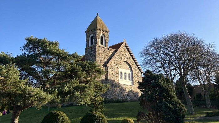 Hyggelig stenkirke med klokketårn omgivet af veltrimmet grønne områder under en klar blå himmel.