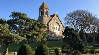 Hyggelig stenkirke med klokketårn omgivet af veltrimmet grønne områder under en klar blå himmel.