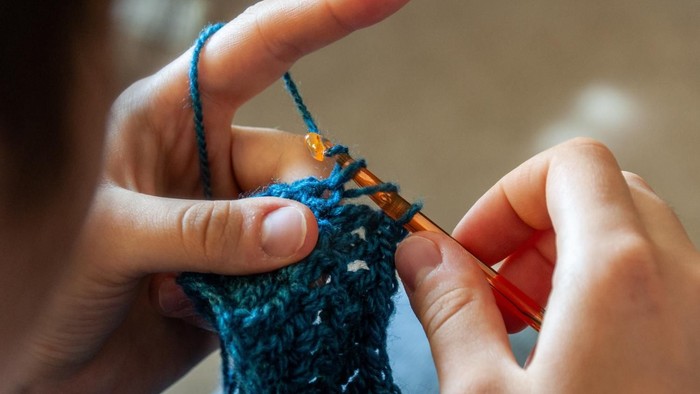 Strickende Hände mit blauem Garn und orangen Stricknadeln.