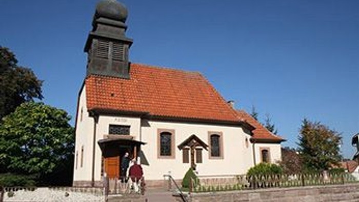 Kleine Kirche mit roter Ziegeldach und Uhrturm, umgeben von Bäumen und Büschen.