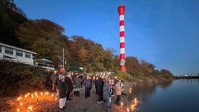 Gruppe von Menschen versammelt sich in der Nähe von Wasser mit brennenden Fackeln, im Hintergrund ein hoher roter und weißer Turm.