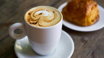 Ein Cappuccino in einer Tasse mit Milchschaumherz und ein Croissant auf einem Teller.