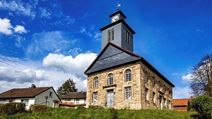 Steinerne Kirche mit Uhrturm in grünem Feld unter blauem Himmel.