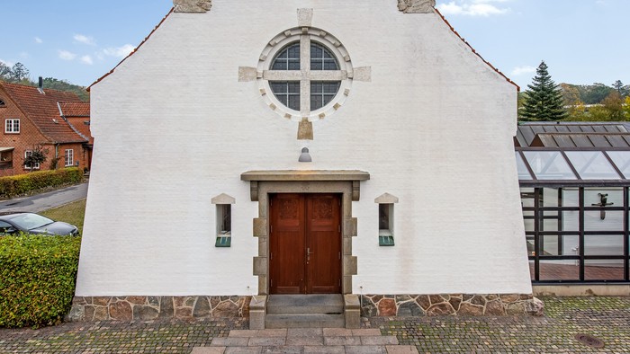 Et hvidt kapel med rund vindueskår og trædør, omgivet af græs og en sti.