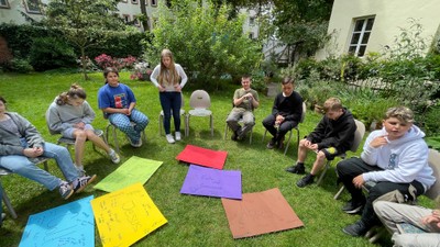 Gruppe von Menschen, die draußen auf dem Gras sitzen und an einer bunten Gruppenaktivität mit großen Papierbögen teilnehmen.