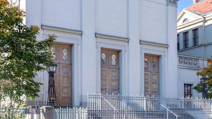 Das Bild zeigt eine weiße Kirche mit Säulen und einem Zaun davor, vor einem klaren blauen Himmel.
