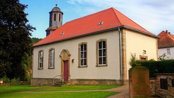 Kleine weiße Kirche mit rotem Dach und Glockenturm, umgeben von grünem Rasen und Bäumen.
