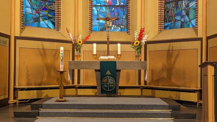 Jahreskerze, links stehend, vor Altar mit zwei Kerzen, zwei Sträußen in Vasen und mittig ein Kruzifix darauf. im Hintergrund drei Kirchenfensterausschnitte.