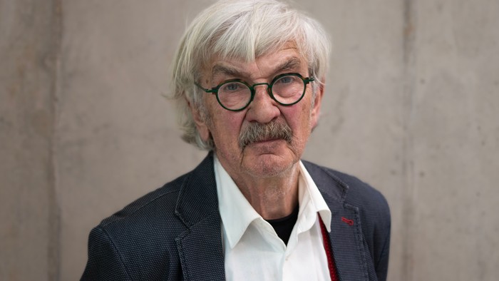 Ein älterer Mann mit weißem Haar und Brille trägt einen dunklen Anzug und eine rote Krawatte.