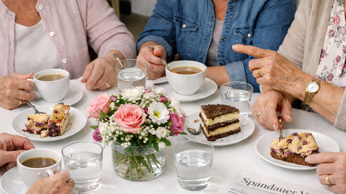 Eine Gruppe genießt Tee, Kuchen und Gespräche an einem Tisch mit Blumen und einer Zeitung.