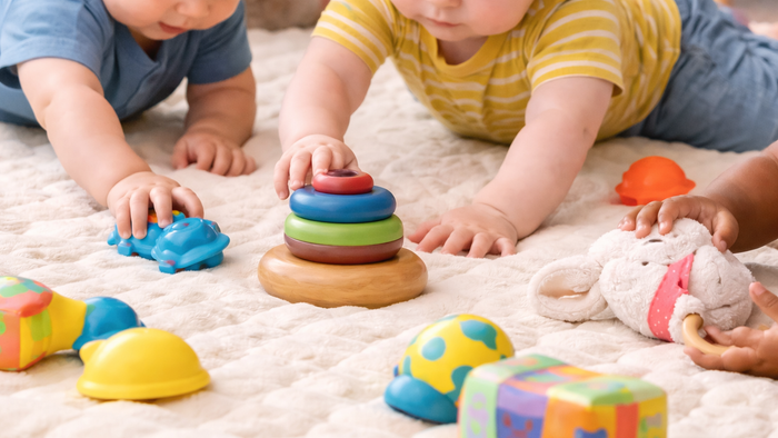Zwei Kleinkinder spielen mit bunten Stapelringen, Bauklötzen und weichen Spielzeugen auf einer weichen Spielmatte.