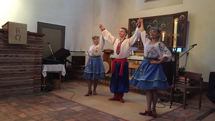 Drei Personen in traditioneller Kleidung tanzen in einem Raum mit Klavier und Kirchenfenster.