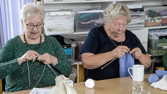 To oldere kvinder sitter ved bordet og stråler med strå. Der er teglampe og tøj på hylderne.