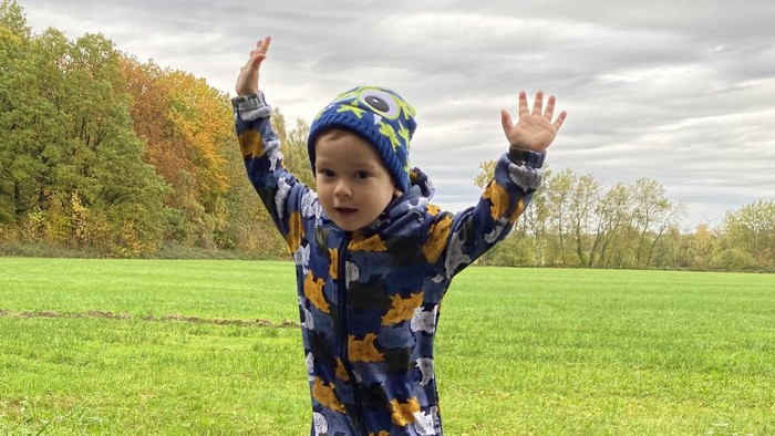 Ein fröhliches Kleinkind in einem Overall mit Blattmuster springt glücklich in eine matschige Pfütze draußen.