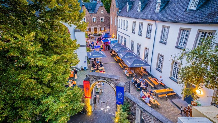Ein malerisches Dorf mit Kopfsteinpflasterstraße, umgeben von historischen Gebäuden und üppigen Bäumen, mit Menschen, die in einem Freiluftrestaurant sitzen.