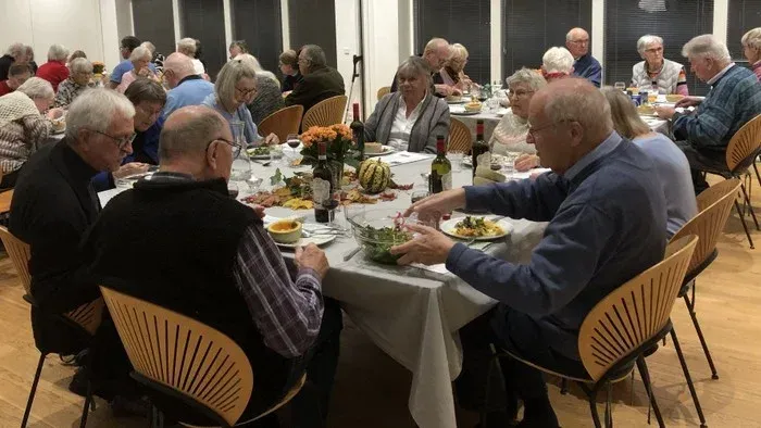 En gruppe ældre folk spiser sammen ved lange bord i en lys og rummelig lokalitet.
