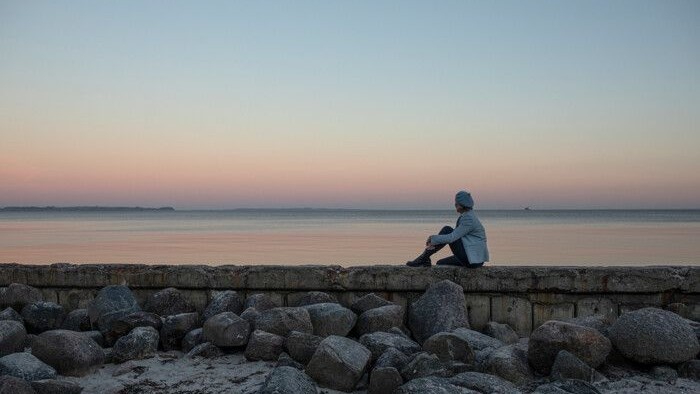 En person sidder på en mur ved stranden og ser ud over havet ved solopgang.
