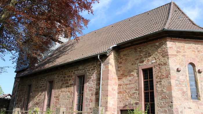 Steinerne Kirche mit Rundbogenfenstern und steilem Dach, umgeben von Bäumen und Gras.