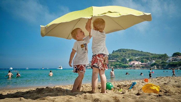 Zwei Kinder stehen am Strand unter einem gelben Regenschirm.