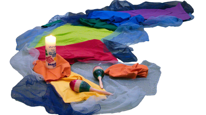 Auf einem bunten Tuch steht eine brennende Kerze. Daneben liegen zwei Rasseln und weitere bunte Tücher.