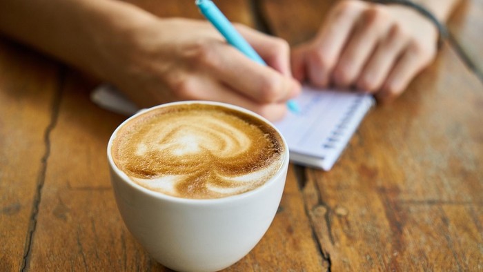Eine Person schreibt in ein Notizbuch, während sie einen Latte-Art-Kaffeebecher mit einem Stift in der Hand hält.