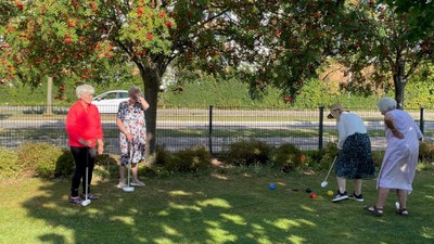 En gruppe ældre spiller udendørs golf med rollatorer på en solrig dag.