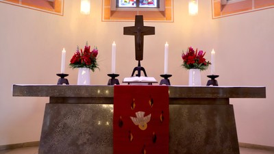 Eleganter Altar mit einem Kreuz, Kerzen, Blumengestecken und einem roten Tuch mit einem Taubenemblem.