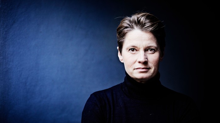 Et alvorligt portræt af en kvinde i en mørk halsturtleneck foran en stemningsfuld blå baggrund.
