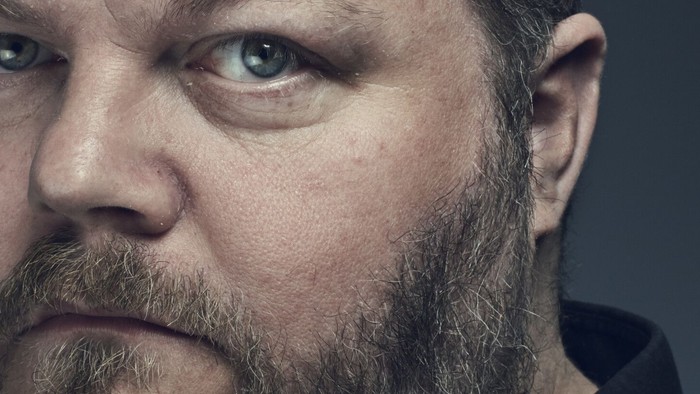 Et nærbillede af en skægget mand med et alvorligt, intenst udtryk mod en mørk baggrund.