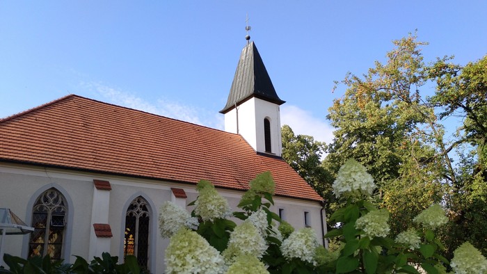 Weiße Kirche mit hohem Turm und roter Ziegeldach, umgeben von üppigen weißen Blumen und grünen Büschen