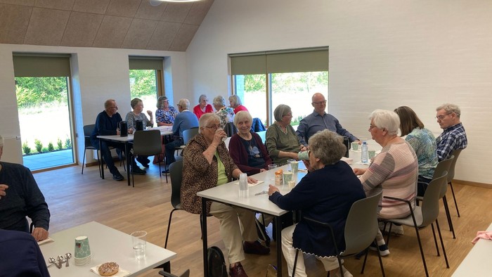 En gruppe ældre personer sidder ved bord i et lys og rummeligt lokal.