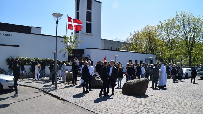 En gruppe mennesker står udenfor et hvidt bygning med dansk flag på en klar dag