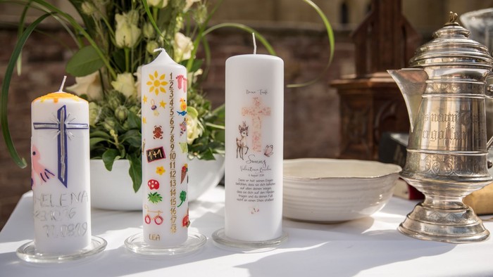 Drei verzierte Taufkerzen stehen auf einem weißen Tischtuch neben einer silbernen Taufkanne, einer weißen Schale und einem Blumenstrauß.