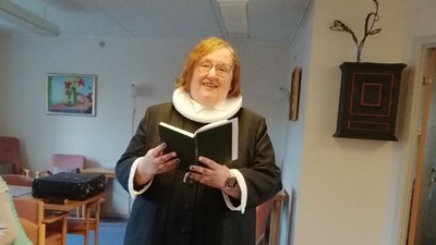 En kvinde i mørkt tøj og hvid krage står og læser fra en bog i et rum med en anden kvinde i baggrunden.