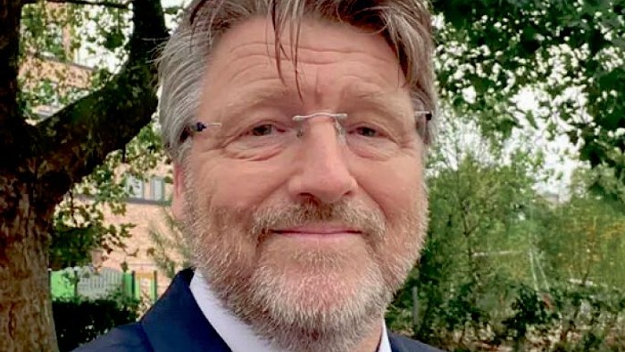 En mand med grå hår og skæg poserer i en have.