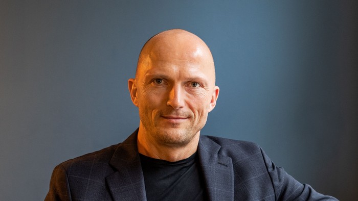 Mand i mørk suite sidder i stue med blå væg.
